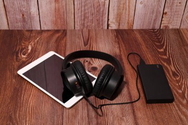 Beyaz bir tabletin üzerinde kablosuz siyah kulaklıklar ve tahta bir yüzey üzerinde taşınabilir bir şarj aleti, müzik dinleme ve iş verimliliği için modern cihazlar bulunmaktadır.