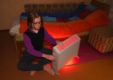 Gözlüklü genç kız yerde oturuyor ve kırmızı bir ısıtıcıyla beyaz elektrikli sobanın düğmesini açıyor.