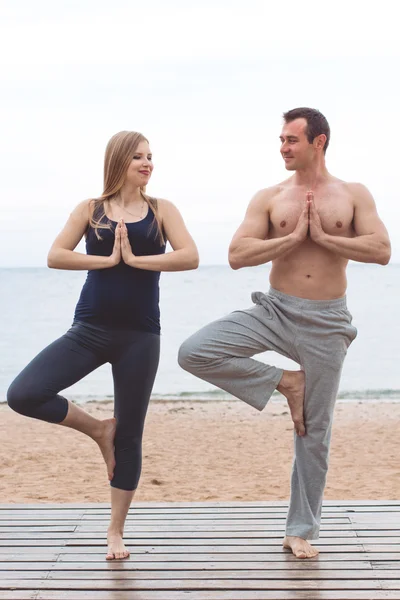 Hombre y mujer embarazada están haciendo yoga en la playa —  Fotos de Stock