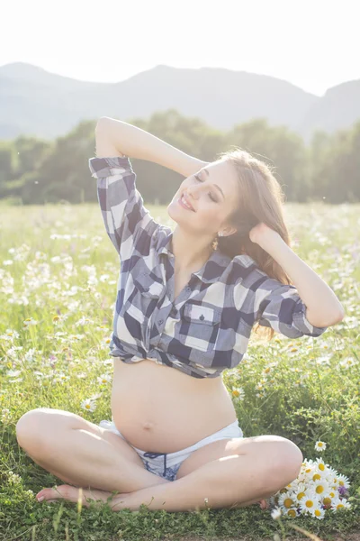 Pregnant woman sitting on nature behind sunset lights — Stock Photo, Image