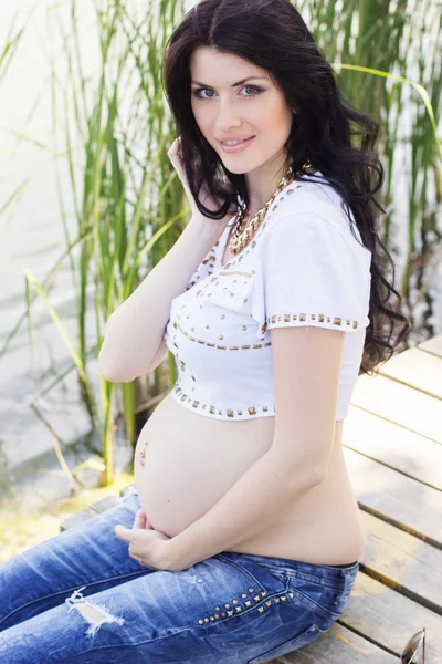 Zwangere brunette meisje, zittend op de brug in de buurt van de rivier — Stockfoto