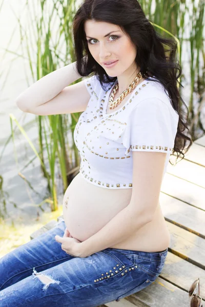 Pregnant brunette woman sitting on bridge near river — Stock Photo, Image