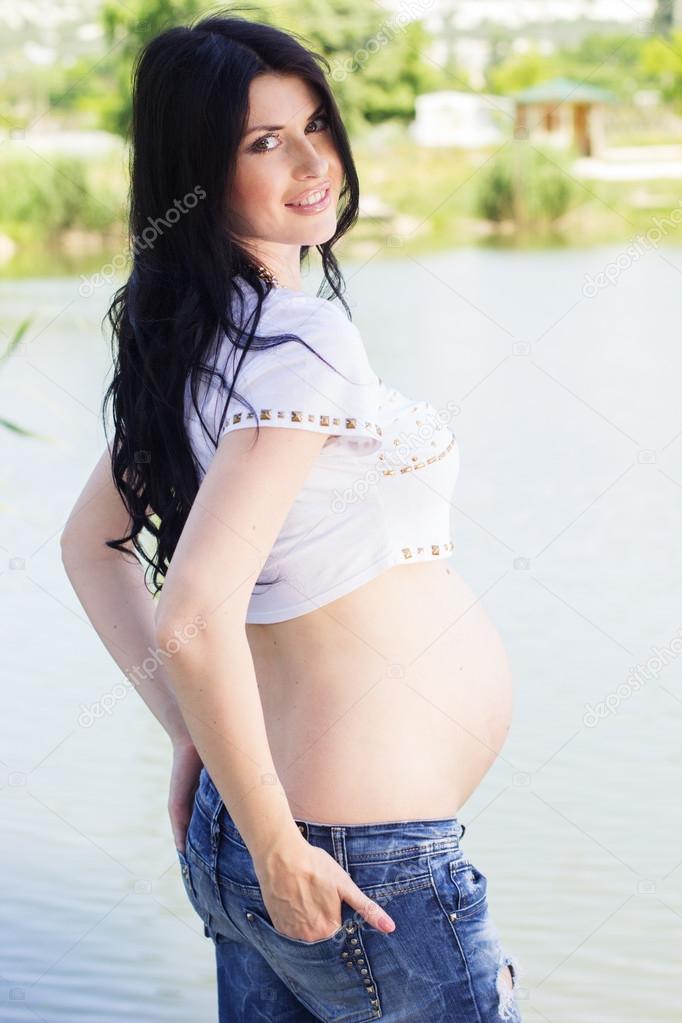 Pregnant brunette girl is standing near blue river