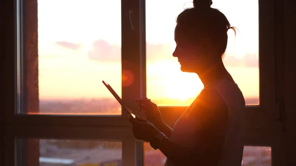 Businesswoman using tablet with sunbeams and lens flare effects against sunset sky window on blurred city background.