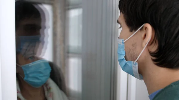 Sad Woman Wearing A Protective Mask Standing At The Door. Mother in medical mask communicated with her adult son through the glass window on the door. Meetings with family. Social distance