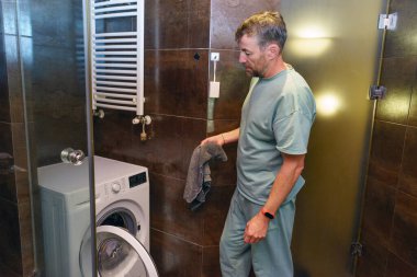 Açık yeşil eşofmanlı orta yaşlı bir adam, elinde bir havluyla modern bir banyoda çamaşır yıkamaya hazırlanıyor..