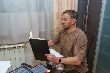 Odaklanmış orta yaşlı bir adam cam bir masada oturup açık siyah bir eskiz defterini inceliyor. Çalışma alanı renkli kalemler ve çizim için boş kağıtlar içeriyor..