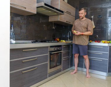 Orta yaşlı bir adam modern bir mutfakta çıplak ayakla yeşil bir elma hazırlıyor. Kahverengi bir tişört ve şort giyiyor. Mutfakta karanlık dolaplar ve paslanmaz çelik aletler var..