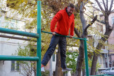 Turuncu ceketli ve gri pantolonlu orta yaşlı bir adam sonbaharda şehir parkında yeşil bir yatay barda kaslarını geliştirir..