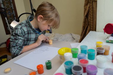 Masadaki beyaz kağıda sarı saçlı odaklanmış genç bir çocuk. Ekose gömlek giyiyor ve yanında renkli guaj kavanozları olan bir fırça kullanıyor..