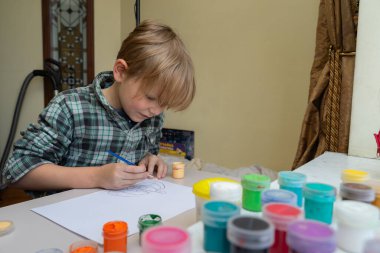 Odaklanmış sarı saçlı genç bir çocuk masada mavi boya fırçasıyla beyaz kağıda resim çiziyor. Ekose gömlek giyiyor ve etrafı boya kavanozlarıyla çevrili..