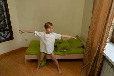 Beyaz tişörtlü genç bir çocuk yatak odasının ahşap zemininde yalınayak duruyor. O, yeşil çarşaflı yapılmamış yatağının yanına uzanmış kollarını uzatarak esniyor. Doğal ışık yakındaki bir pencereden odayı dolduruyor..