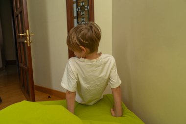 Arka planda, sarı saçlı, yeşil yastıklı genç bir çocuk var. Krem rengi bir tişört giyiyor ve sola bakıyor. Duvardaki alanı kopyala.