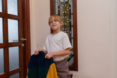 Genç sarışın bir çocuk koridorda askısında renkli yün bir ceket tutuyor. Evde giyinirken yüzünü buruşturuyor..