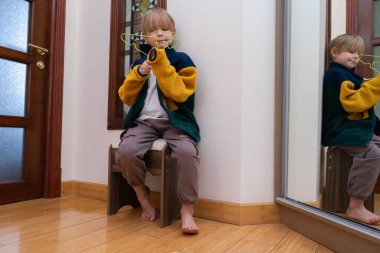 Kapalı bir taburede oturan genç bir çocuk, elinde yeşil bir askıyla. Renkli bir yün ceket giyiyor ve yalınayak. Bir ayna onun görüntüsünü yansıtır..