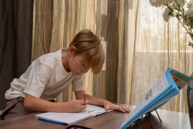 Beyaz tişörtlü, odaklanmış sarışın bir çocuk masadaki bir deftere yazıyor. Çizgili perdelere karşı açık bir kitapla ödev yapıyor..