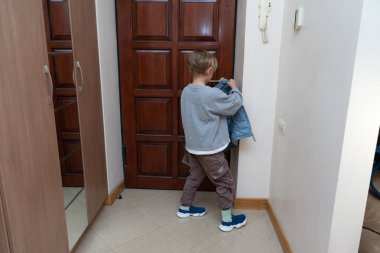 Genç bir çocuk koridorda duruyor ve ahşap bir kapının yanında mavi bir ceket giyiyor. Gri bir kazak ve kargo pantolonu giyiyor, evden çıkmaya hazırlanıyor..