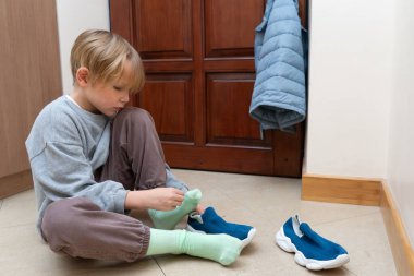 Genç bir çocuk kapalı bir yerde fayanslı bir zeminde oturuyor, yeşil çoraplar giyiyor. Mavi spor ayakkabılar ve asılı bir ceket ahşap bir kapının yanında görünür..