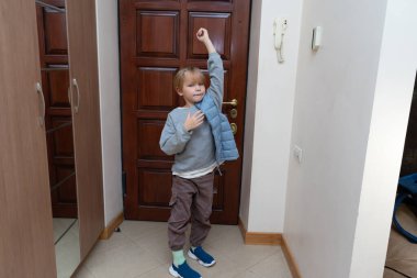 Genç sarışın bir çocuk evin girişinde duruyor, gri bir kazağın üzerine açık mavi bir kabarık yelek giyiyor. Dışarı çıkmaya hazırlanıyor..