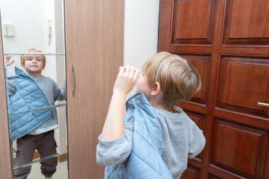 Sarışın genç bir çocuk ev koridorunda giyiniyor, mavi örtülü bir yelek giyiyor ve aynalı gardırop kapısındaki yansımasına bakıyor..