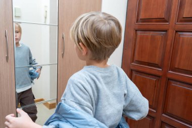 Sarı saçlı, arkadan görülen genç bir çocuk, koridorda aynalı bir gardırop kapısındaki yansımasına bakarken mavi bir ceket giyer..