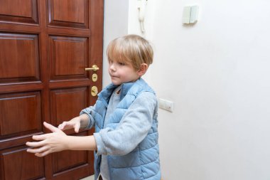 Mavi yelek ve gri gömlek giyen genç sarışın bir çocuk kahverengi ahşap bir kapının yanındaki ev girişinde duruyor. O, ellerini uzatarak yana bakıyor..