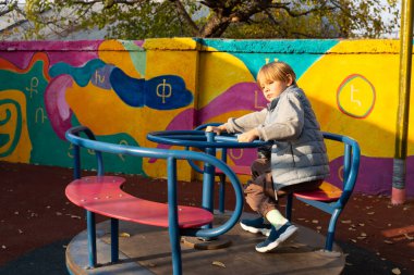 Sarı saçlı genç bir çocuk oyun parkında renkli bir atlıkarıncada yalnız oturuyor. O bir yelek giyiyor ve dalgın bir bakışı var. Arkasında soyut şekilleri ve Ermeni harfleri olan canlı bir duvar resmi var. Sahne sıcak güneş ışığıyla aydınlanıyor.