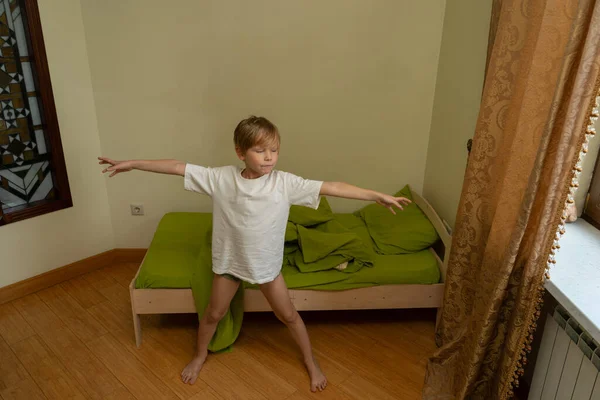 Beyaz tişörtlü genç bir çocuk yatak odasının ahşap zemininde yalınayak duruyor. O, yeşil çarşaflı yapılmamış yatağının yanına uzanmış kollarını uzatarak esniyor. Doğal ışık yakındaki bir pencereden odayı dolduruyor..