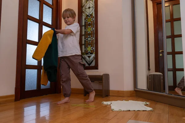 Genç bir çocuk ahşap bir zeminde çıplak ayakla yün bir ceket giyiyor. Cam kapılı bir koridorda yerde bir tişört ve askı var..