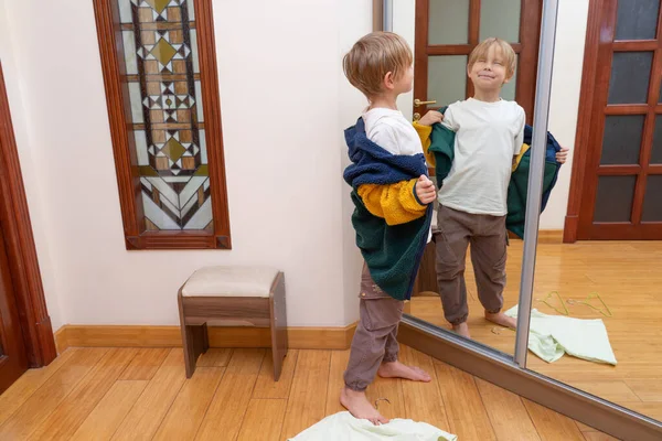 Komik sarışın bir çocuk aynanın önünde çıplak ayakla duruyor, dağınık bir odada aptal bir yüz ifadesi takınırken renkli bir yün ceket giymeye çalışıyor..