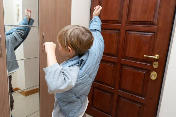 Arkadan görülen genç bir çocuk gri bir gömleğin üzerine açık mavi yorgan giymişti. Evinin koridorunda, ahşap bir kapı ve hareketlerini yansıtan aynalı bir gardırobun yanında duruyor..