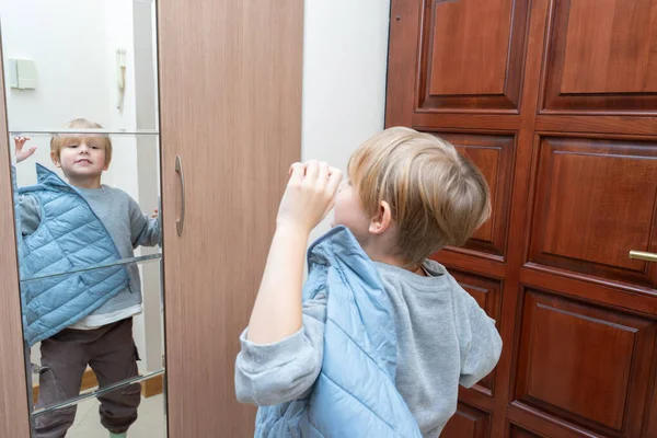 Sarışın genç bir çocuk ev koridorunda giyiniyor, mavi örtülü bir yelek giyiyor ve aynalı gardırop kapısındaki yansımasına bakıyor..