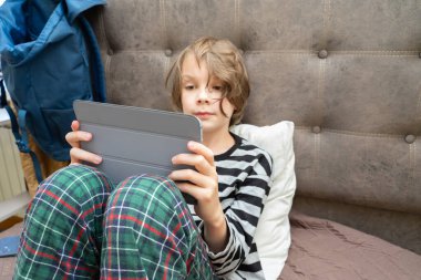 Çizgili pijamalı genç bir çocuk elinde tuttuğu dijital tablete odaklanmış bir şekilde yatakta oturuyor. Bir yastığa ve kahverengi bir yatak başlığına yaslanıyor..