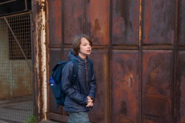 Sırt çantalı genç bir çocuk büyük, paslı endüstriyel metal bir kapının önünden geçiyor. Mavi bir ceket giyiyor ve düşünceli bir ifadesi var. Şehir ortamının cesur ve yıpranmış bir havası var..