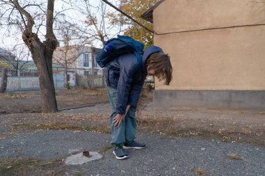 A boy wearing a blue jacket and a backpack bends over, holding his knees on an asphalt path. The outdoor setting suggests an autumn day in a yard.