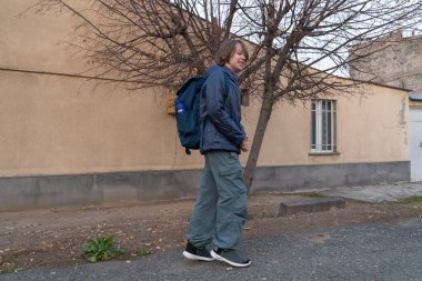 Büyük mavi bir sırt çantası olan gülümseyen ergen bir çocuk şehir yolunda yürüyor. Mavi bir ceket ve kargo pantolonu giyiyor. Sonbaharda serin ve bulutlu bir günde okula gidip gelmeyi öneriyor..