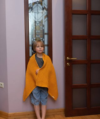 Genç bir çocuk, rahat sarı bir waffle örgü battaniyeye sarılı bir kapının yanında duruyor. Evin koridorunda rahat görünüyor..