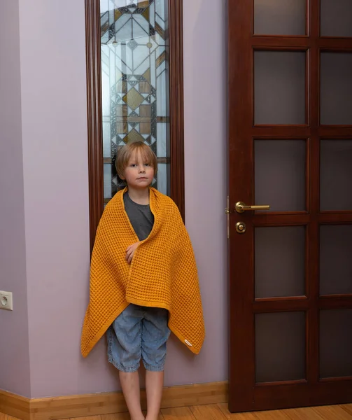 Genç bir çocuk, rahat sarı bir waffle örgü battaniyeye sarılı bir kapının yanında duruyor. Evin koridorunda rahat görünüyor..