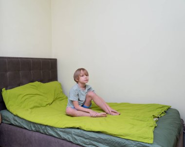 Sarışın, beyaz bir çocuk yeşil yorganı olan bir yatakta yalınayak oturuyor. Açık yeşil bir tişört ve mavi bir şort giyiyor. Direkt olarak kameraya tarafsız bir ifadeyle bakıyor. Odanın düz krem duvarları ve kahverengi tüyleri var.