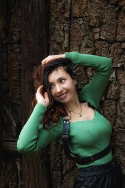 Dokulu ahşap kalasların önünde duran bir kadının açık hava portresi. Onun yeşil kazağı ve siyah deri şortu doğal ortama kontrastlık ekler. Kendine güvenen, rahat bir duruş güçlü bir moda ve yaşam tarzı estetiği yaratır..