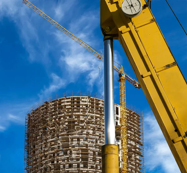 Part of a construction machine (excavator or crane) with multi-storey building under construction with scaffolding (new residential complex) on the background, Moscow, Russia