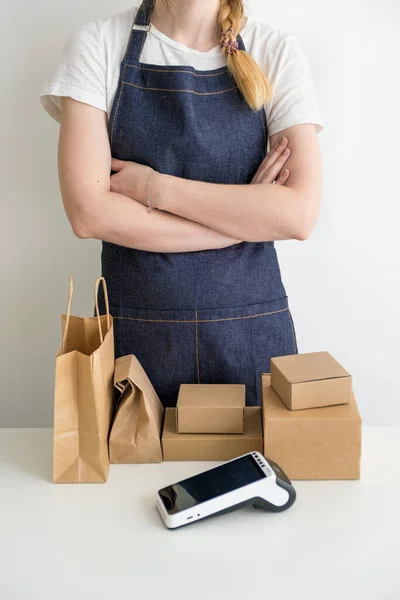 Jeune femme portant un tablier avec les bras croisés avec des boîtes en carton, emballages en carton et terminal de paiement moderne à la table — Photo