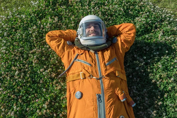 Happy cosmonaut wearing orange spacesuit having a rest while lying on green grass — Stock fotografie