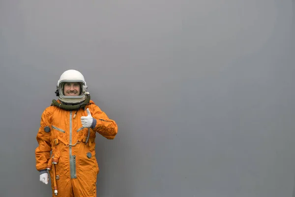 Šťastný astronaut v oranžovém skafandru a helmě ukazující palce nahoru gesto na šedé zdi pozadí — Stock fotografie