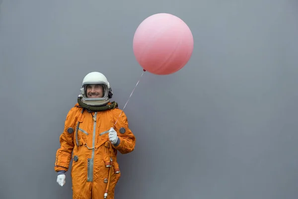 Astronaut na sobě oranžový kosmický oblek a helma drží růžový nafukovací balón proti šedé zdi pozadí — Stock fotografie