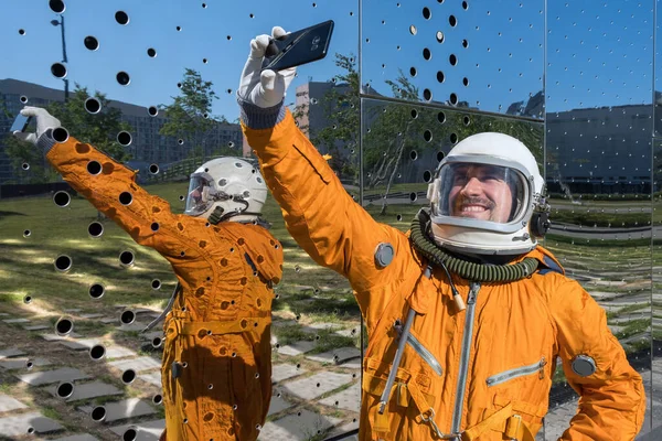 Gelukkig astronaut dragen oranje ruimtepak en ruimte helm houden mobiele telefoon en het nemen van selfie buiten. — Stockfoto