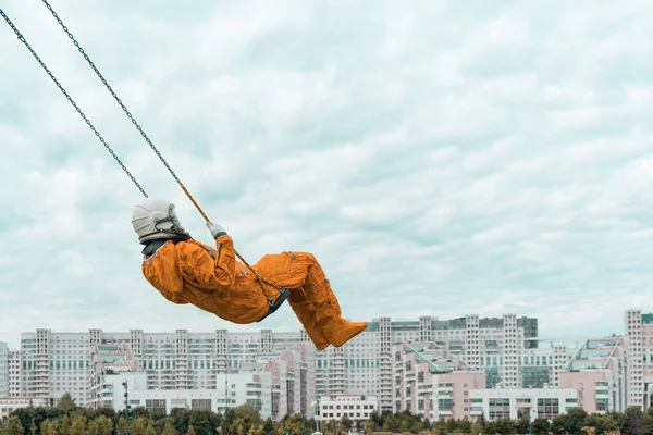 Astronaut v oranžovém skafandru a helmě na houpačce proti zatažené obloze venku — Stock fotografie