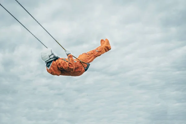 Astronaut in orangefarbenem Raumanzug und Weltraumhelm auf Schaukel gegen bewölkten Himmel — Stockfoto