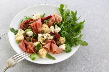 Salata roka, güneşte kurutulmuş domatesli Parma jambonu, mozzarella dilimleri, kızarmış ekmek, kapari, baharat