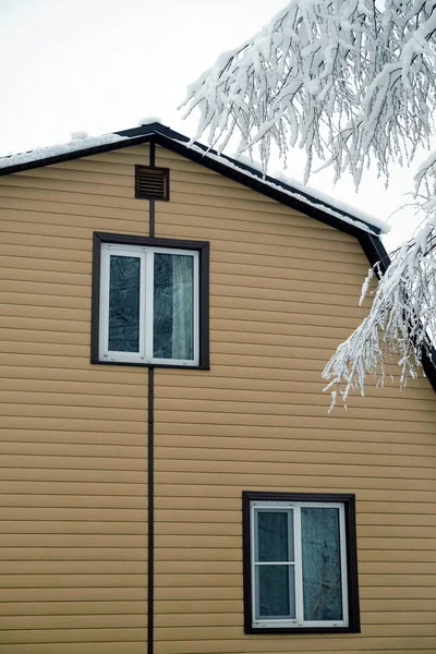 カントリーハウスの壁にプラスチック製の窓がある上階には ベージュのサイドと雪の表面と木々の枝で覆われた茶色の金属屋根で覆われています — ストック写真